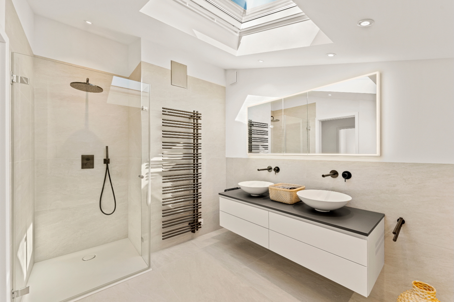 Modern bathroom in a renovated detached house in Zürich with walk-in glass shower, rain showerhead, heated towel rail, double vessel sinks on floating vanity, and skylight.