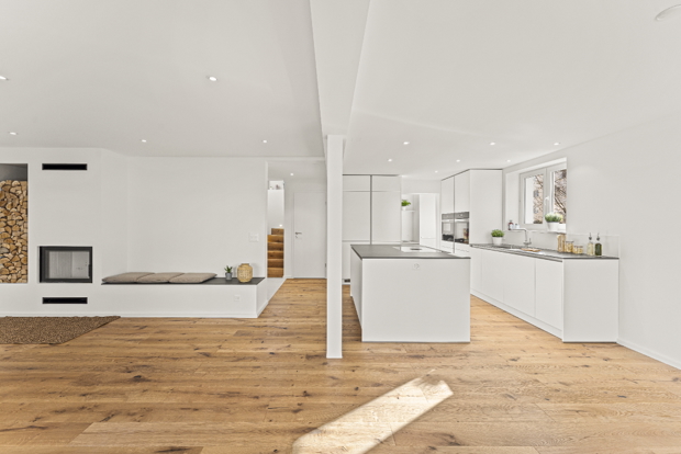 Spacious open-plan living and kitchen area in a renovated detached house in Zürich with white kitchen island, built-in fireplace with wood storage, oak hardwood flooring, and recessed lighting.