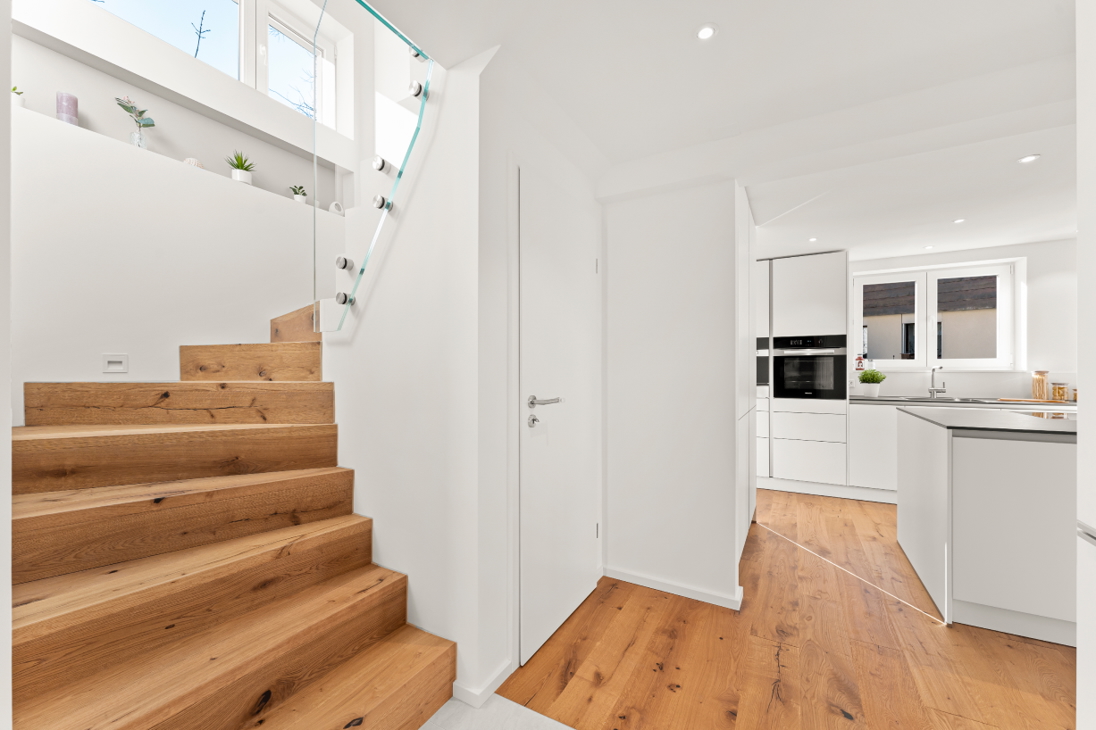 Oak staircase with frameless glass railing in a renovated detached house in Zürich featuring decorative shelf niche with plants and view towards kitchen.